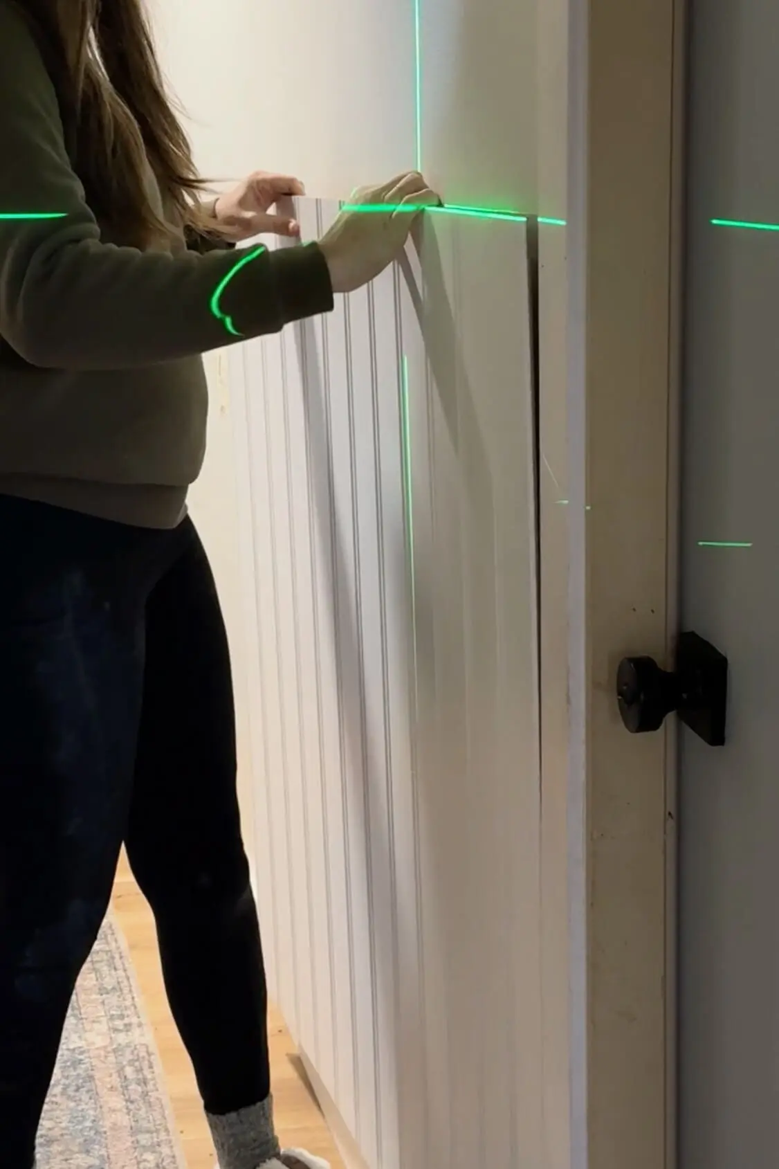 Close-up of a DIY beadboard installation project showing hands carefully aligning a vertical panel against a wall. A bright green laser level projects horizontal and vertical guide lines to ensure precision in the placement.